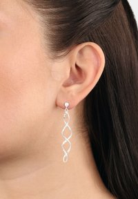 Silver snurriga hängande örhängen med en oändlighetsloop-design bar på höger öra med mörkbrunt hår.