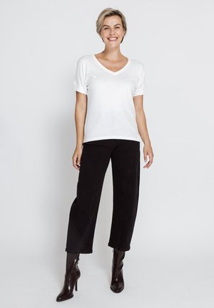 Smiling woman standing, wearing white V-neck T-shirt, black wide-leg pants, and dark brown heeled ankle boots against plain background.