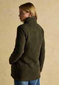 Kobieta z blond włosami, ubrana w ciemnooliwkową polarową kurtkę z wysokim kołnierzem i niebieskie dżinsy, stoi na beżowym tle.