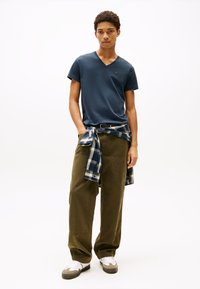 Young man standing with hands in pockets, wearing navy v-neck t-shirt, olive cargo pants, white sneakers, and a plaid shirt tied around waist.