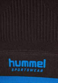 Gros plan sur un tissu noir côtelé avec le texte bleu « "hummel SPORTSWEAR" » et une bande bleue horizontale en bas. Aucune personne visible.