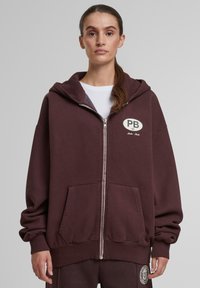 Bordová mikina na zips vyrobená z textúrovanej tkaniny, s bielym kruhovým logom na hrudi, veľkými vreckami a kapucňou.