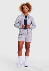 Γυναίκα με σγουρά μαλλιά που φορά γκρι φούτερ με φερμουάρ και σορτς, ναυτικό μπλουζάκι, λευκές κάλτσες και λευκά αθλητικά παπούτσια, στέκεται με τα χέρια της κοντά στο φερμουάρ.