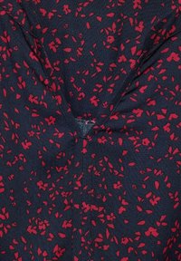 Tela azul oscura con pequeño patrón floral rojo disperso y un detalle de costura en forma de V cerca del centro.