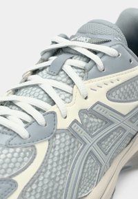Lys grå treningssko med et mesh-øvre, kontrasterende hvite detaljer, teksturerte overlegg og flate hvite skolisser. Merkbar ASICS-merking.