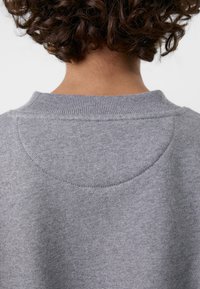 Vue de dos d'une personne aux cheveux bruns bouclés portant un pull en maille gris avec un col côtelé et un détail de couture semi-circulaire.