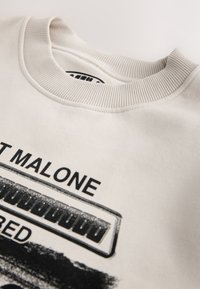 Sweat-shirt beige avec col rond côtelé, avec texte noir "MALONE" et illustration d'un harmonica ou d'un instrument à clavier.