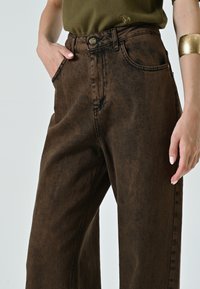 Pantalons en denim marron foncé taille haute avec une texture décolorée, présentant une fermeture à bouton en métal, des passants de ceinture et des poches latérales, stylés avec une coupe décontractée.