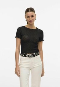 Svart strikket topp med korte ermer, med en bølget hals og teksturert mønster, sammen med hvite jeans og et svart belte med metalldetaljer.