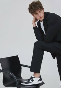 Černobílé tenisky s kulatým akcentem, vyrobené ze směsi hladké a texturované kůže, v kombinaci s elegantním černým oblekem a bílým tričkem.