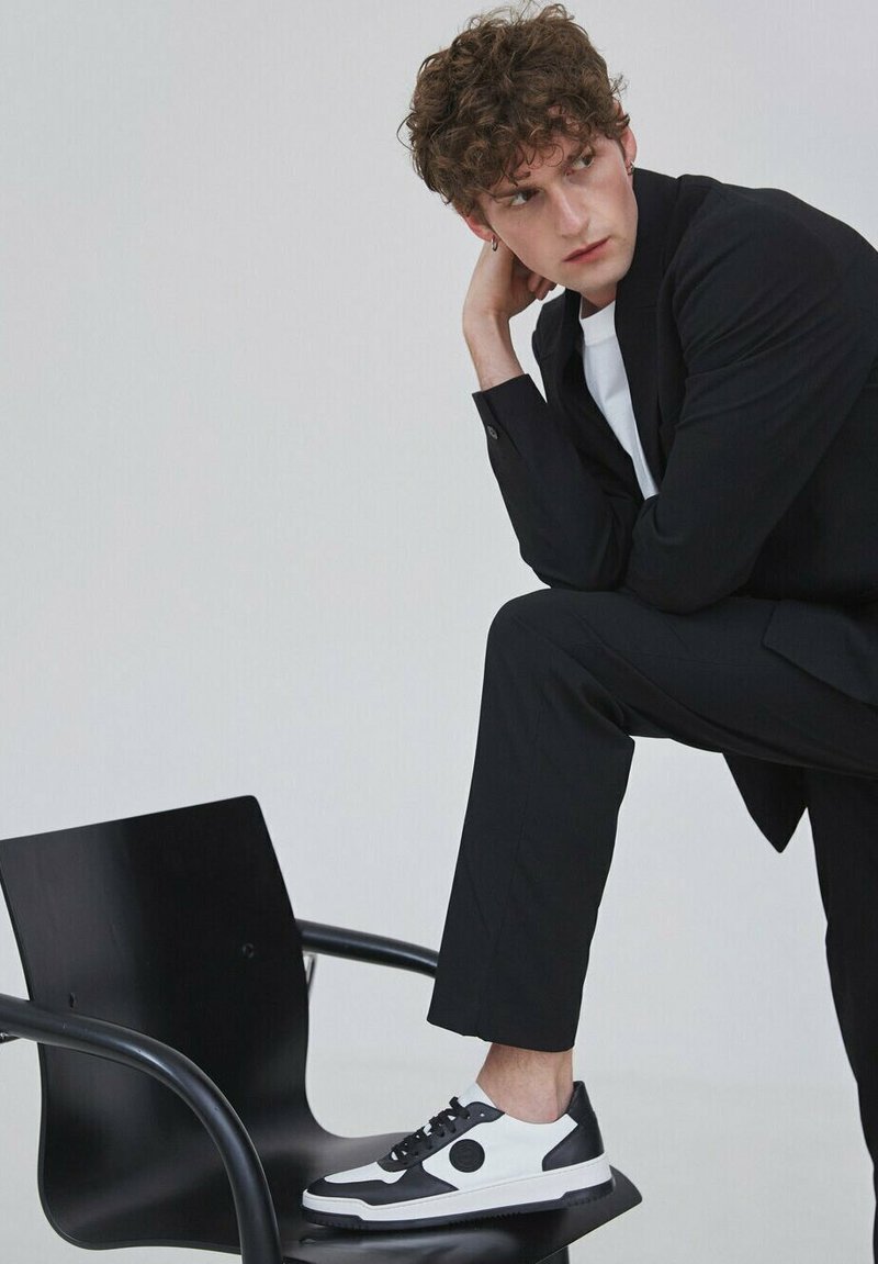 Černobílé tenisky s kulatým akcentem, vyrobené ze směsi hladké a texturované kůže, v kombinaci s elegantním černým oblekem a bílým tričkem.