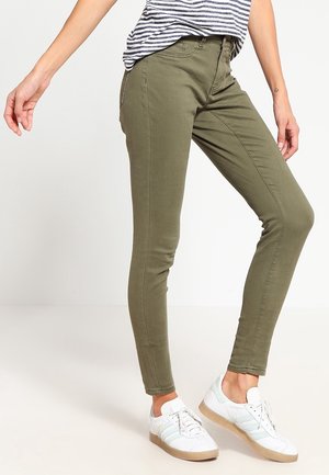 Person, die olivgrüne schmale Hose, weiße Sneakers mit Gummisohlen und ein schwarz-weiß gestreiftes Shirt mit einer sichtbaren Hand trägt.