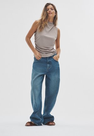 Ärmelloses Tan-Oberteil mit drapiertem Stoff. Hellblaue, locker sitzende Jeans mit verblasster Textur. Braune Slides-Sandalen ergänzen das Outfit.