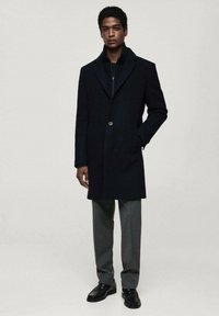 Dark wool overcoat with a notched collar, single button closure, and side pockets; paired with gray trousers and black shoes.