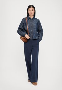 Denim shirt with a patterned texture, navy wide-leg pants, and brown crossbody bag. Model standing against a neutral background.