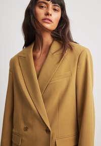 Blazer double boutonnage moutarde en tissu texturé, avec revers, poche poitrine et boutons marron.