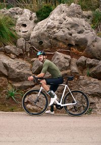 Vélo gravel blanc avec des pneus noirs. Cycliste en maillot olive et short noir, portant un casque noir avec des accents verts. Arrière-plan rocheux.