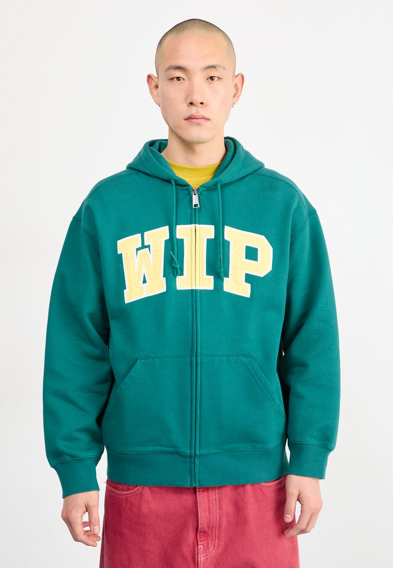 Grønn zip-up hettegenser med gul "WIP" tekst, frontlommer og snorhette. Brukt med en gul skjorte og rosa bukser.