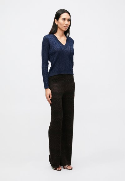 Jūros mėlynos spalvos v formos kaklo megztinis su tekstūriniu zigzago raštu, derinamas su aukštu juosmeniu juodomis kelnėmis su banguotomis linijomis; modelis avintis sandalus.