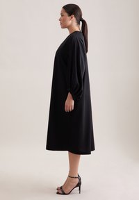 Czarna sukienka do kolan z obszerne długie rękawy, gładka w dotyku i luźnym kroju. Do tego czarne sandały na wysokim obcasie z paskami.