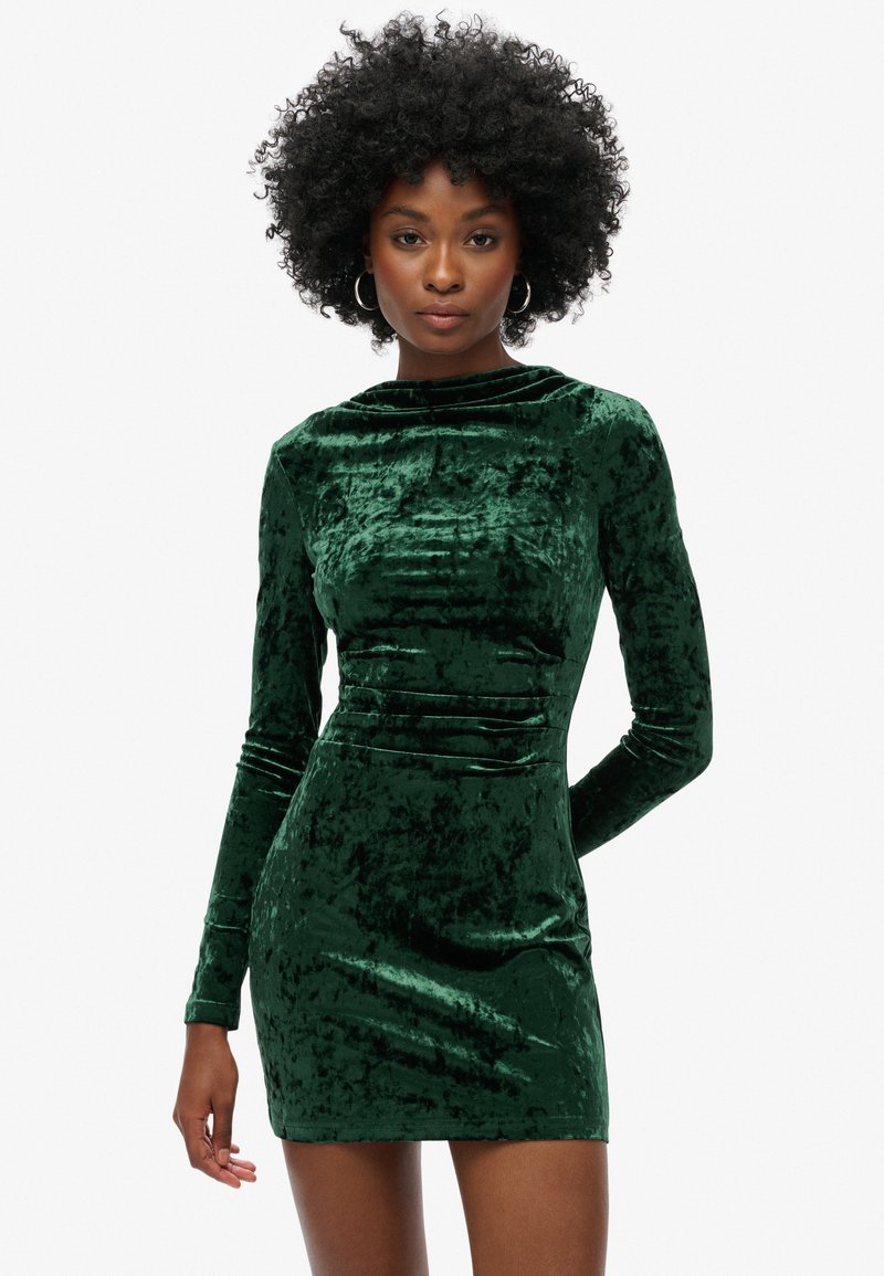 Vestido verde de terciopelo con mangas largas y cuello alto. Diseño ajustado con sutil fruncido, mostrando una superficie suave y texturizada.