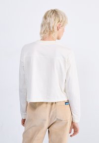 Gekürztes weißes Baumwollhemd mit langen Ärmeln und lockerer Passform. Verfügt über sichtbare Nähte und ein einfaches, minimalistisches Design. Kombiniert mit beigefarbenen Cordhosen.