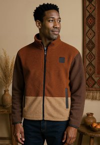 Fleecejack met een kleurblokontwerp in bruin, roest en tan, met een hoge kraag, zwarte rits en zijzakken.