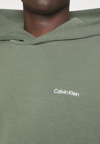 Olivově zelená bavlněná mikina s měkkou texturou, s bílým logem "Calvin Klein" na přední straně a designem kapuce na šňůrku.