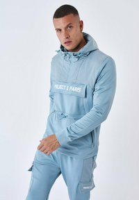 Ljusblå hoodie med dragkedjeficka, dragsko i huvan och matchande byxor. Har vit text "PROJECT X PARIS" på bröstet och byxorna.