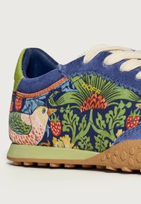 Sneaker con tessuto blu decorato con motivi floreali e uccelli, dettagli verdi, lacci crema e una suola marrone texturizzata.