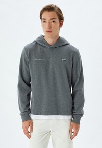 Sudadera con capucha gris y cordón, hecha de una tela texturada. Presenta un pequeño logo en el pecho y letras en el lateral.