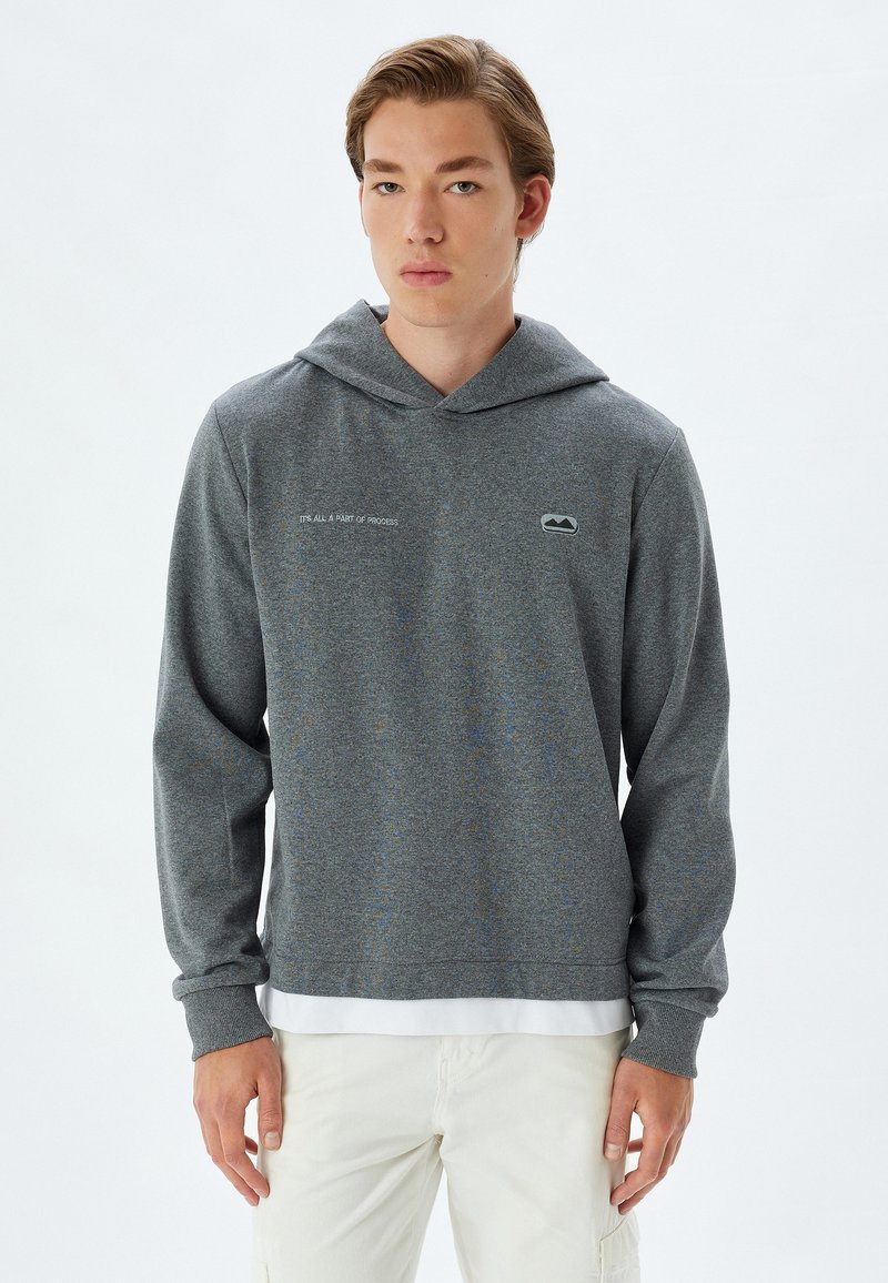 Sudadera con capucha gris y cordón, hecha de una tela texturada. Presenta un pequeño logo en el pecho y letras en el lateral.