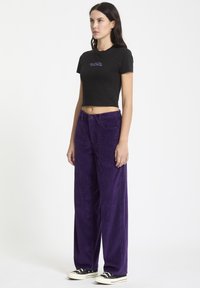 T-shirt noir court avec du texte violet, associé à un pantalon en velours côtelé violet ample et à nervures. Le modèle porte des baskets noires avec des semelles blanches.