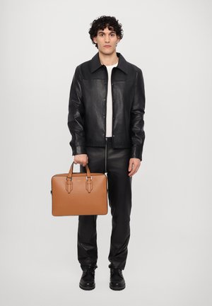 Black leather jacket, white top, black leather pants. Holding a tan briefcase with double handles and buckle accents. Black boots.