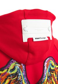 Sudadera con capucha roja con alas de lentejuelas coloridas en la espalda y una etiqueta blanca que dice "MWM Design" adherida a la capucha.