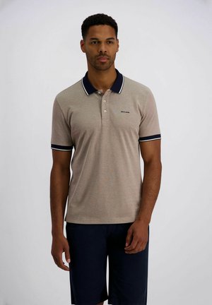 Mann trägt beigefarbenes Polohemd mit marineblauem Kragen und Besatz sowie dunkelblaue Shorts, steht vor weißem Hintergrund.