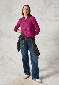 Camicia fucsia con bottoni e motivi geometrici neri, abbinata a jeans larghi in denim scuro e una giacca di denim nera legata in vita. Sneakers bianche completano il look.