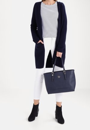 Woman wearing a navy cardigan, striped white and navy shirt, white pants, black ankle boots, holding a navy handbag by the handles.