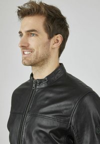 Schwarze Lederjacke mit glatter Textur, mit hohem Kragen und Frontreißverschluss. Steppnähte und Logo am Ärmel sind sichtbar.