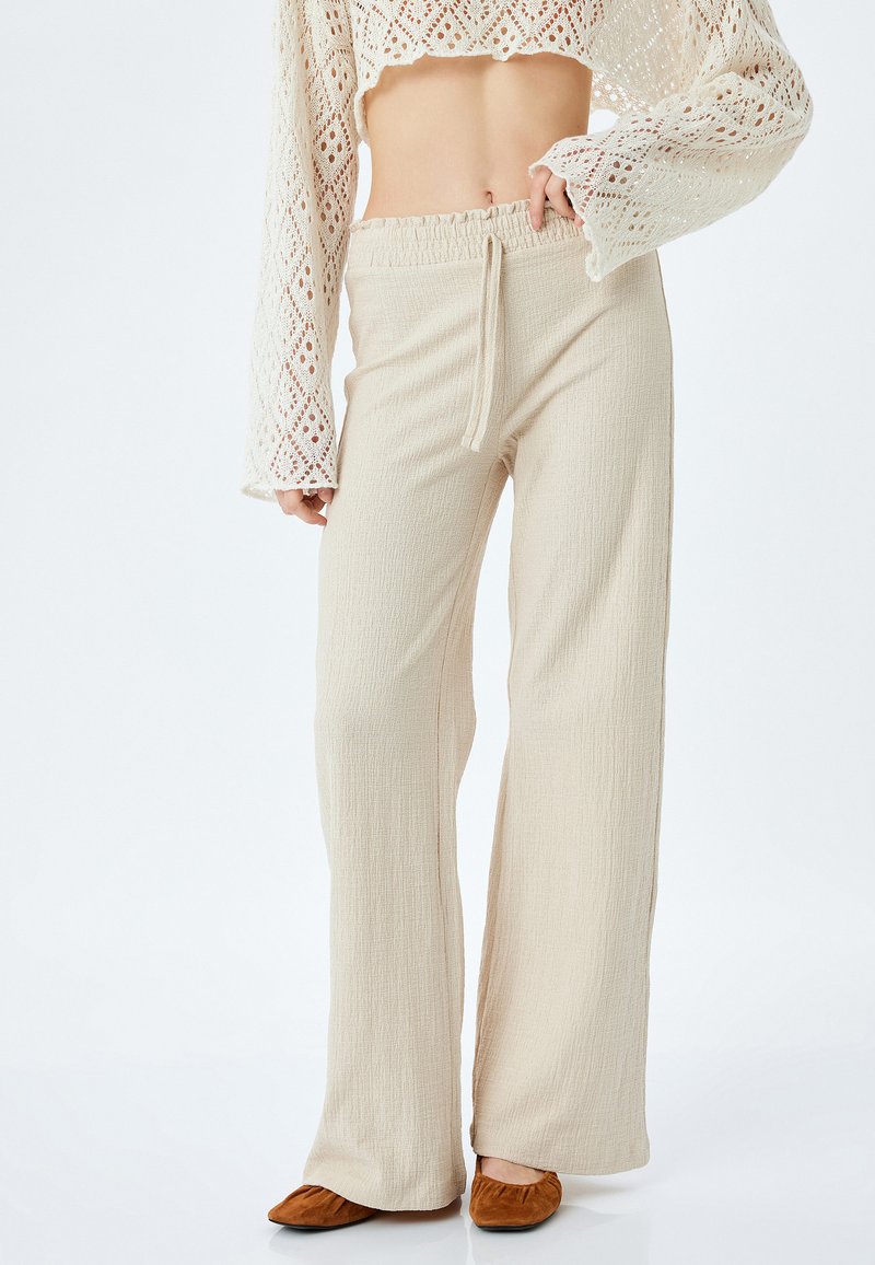 Beige wijde broek met textuurstof, elastische tailleband en trekkoord. Gecombineerd met bruine platte schoenen en een witte kanten crop top.