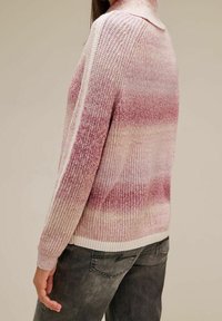 Femme portant un pull en maille dégradé rose et blanc et un jean gris foncé, vue de dos et de côté sur un fond uni.