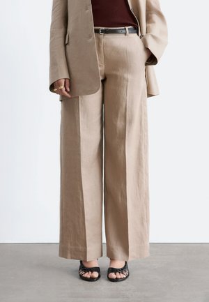 Pantalon beige à jambes larges accompagné d'un blazer assorti, ceinture noire, haut bordeaux, bagues dorées et sandales noires tissées à bout ouvert.