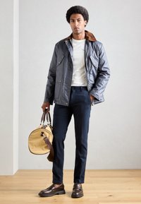 Veste imperméable grise avec col marron, chemise blanche, pantalon marine et mocassins marron. Tenant un sac rond jaune avec des anses marron.