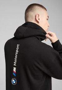 Hoodie noir avec un col montant. Présente un texte blanc "Motorsport" et le logo BMW sur le côté. Tissu doux avec une texture lisse.