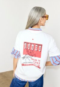 Γυναίκα με γκρίζα μαλλιά φοράει λευκό μπλουζάκι με κόκκινο σχέδιο ροντέο και ριγέ πουκάμισο από κάτω, στέκεται μπροστά σε ένα απλό φόντο.