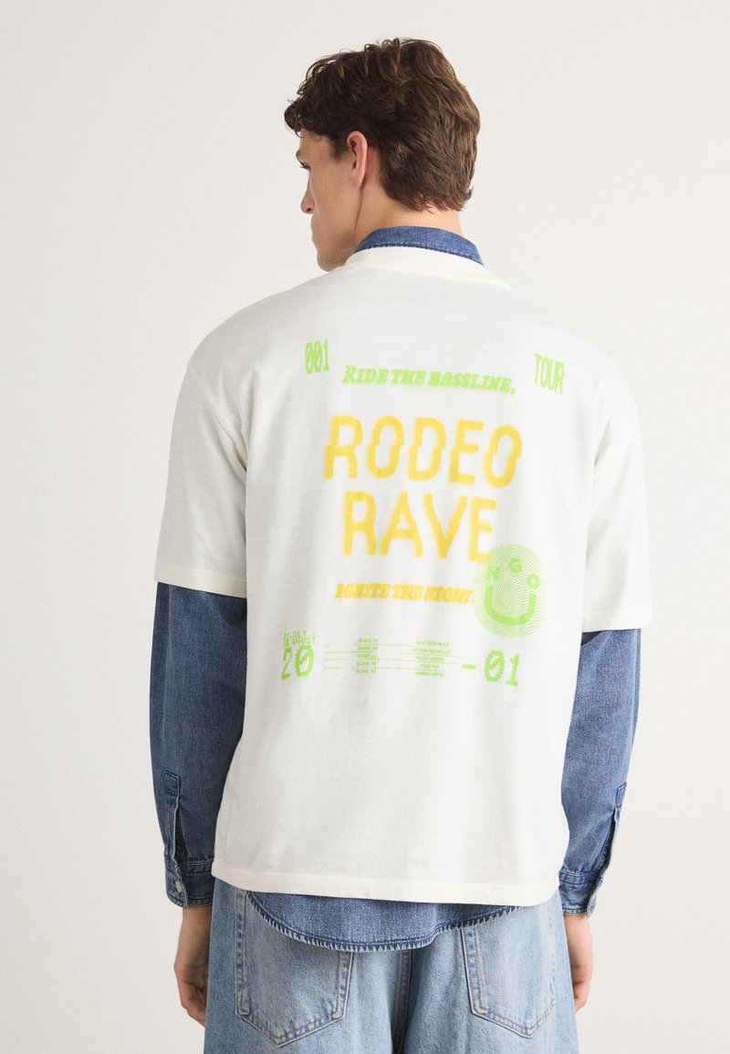 Biała koszulka z krótkim rękawem z kolorowym nadrukiem z tyłu. Grafika zawiera napis "RODEO RAVE" z żółtymi i zielonymi akcentami.