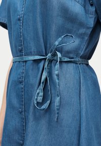 Τζιν φόρεμα με υφασμάτινη ζώνη στη μέση δεμένη σε φιόγκο, λεπτομέρειες με κουμπιά κατά μήκος του μπροστινού μέρους και κοντά μανίκια σε ένα άτομο.