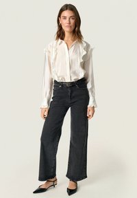 Hvid, rynket bluse med lange ærmer, parret med mørke, højtaljede jeans; sorte spidse slingback-sko fuldender outfittet.