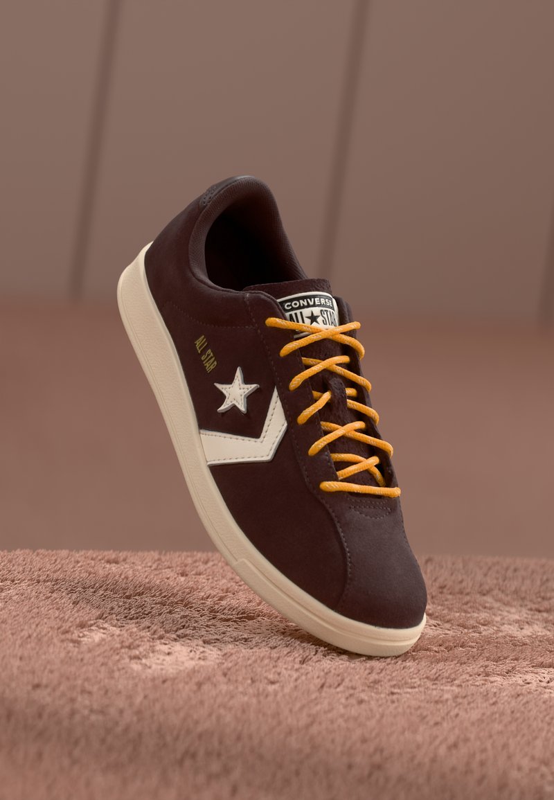 Zapatillas de ante marrón con logotipo de estrella blanca, acentos laterales blancos y cordones naranjas. Presentan una suela de goma texturizada y un cuello acolchado.