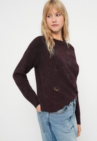 Burgundy knitted sweater with long sleeves, round neckline, featuring heart patterns and a distressed detail near the hem. Mixed texture.